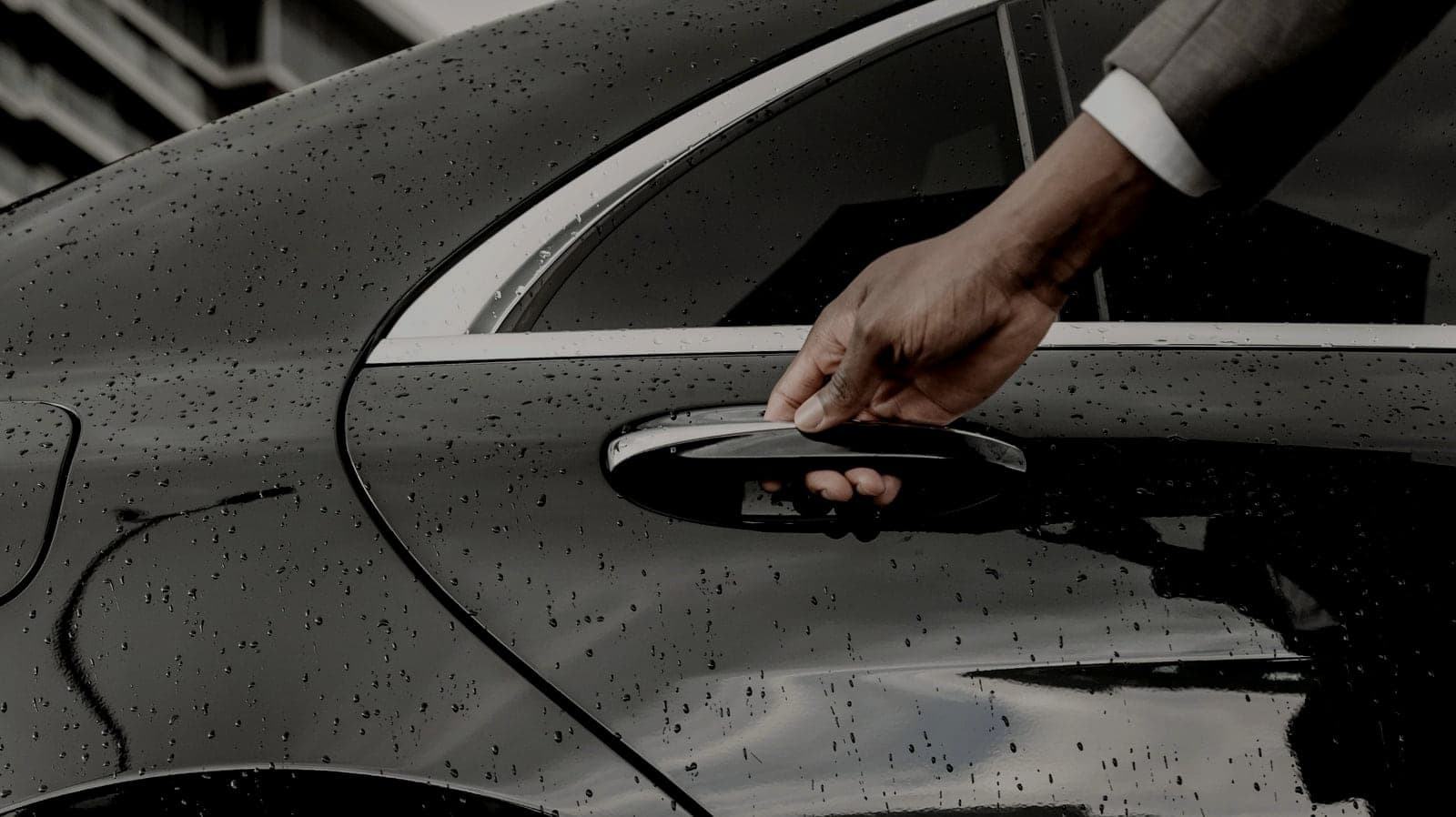 A chauffeur's hand opening the rear door of a black sedan in the rain